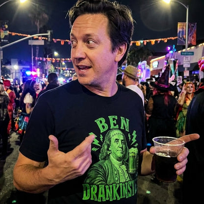 A man wearing a dark blue Ben Drankinstein t-shirt at a crowded nighttime street party. The green graphic shows Benjamin Franklin as Frankenstein holding a beer mug.