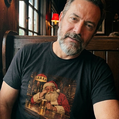 A bearded man sitting in a wooden pub booth wearing a black t-shirt featuring a vintage graphic of Santa Claus drinking beer at a bar.