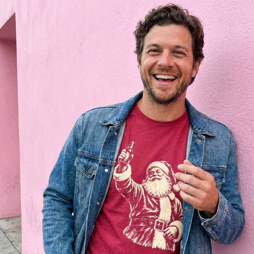 A man wears a red t-shirt with a vintage Santa Claus drinking beer graphic and a denim jacket, posing against a bright pink wall.