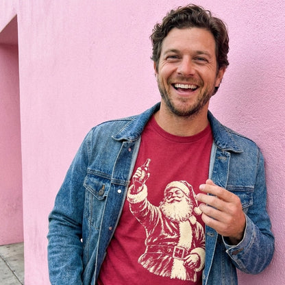 A man wears a red t-shirt with a vintage Santa Claus drinking beer graphic and a denim jacket, posing against a bright pink wall.