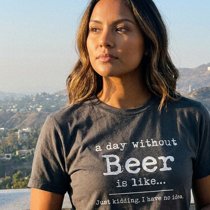 A woman stands on a rooftop overlooking the Hollywood Hills wearing a dark grey t-shirt that says 'a day without Beer is like... Just kidding, I have no idea.'